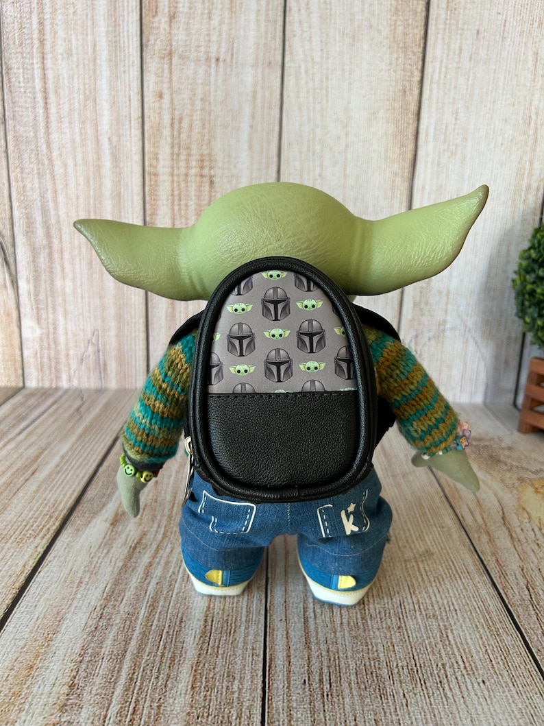 May include: A green-skinned creature figure with large ears wearing a striped knit sweater, blue denim pants with white stitching, and a small black backpack featuring a repeating pattern of grey helmets and green creature faces on the back.