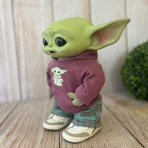 Op de afbeelding: Een groen wezenfiguurtje met grote donkere ogen en puntige oren, gekleed in een mauve hoodie met een kleine afbeelding van hetzelfde wezen op de voorkant, gecombineerd met een groen-zwarte geruite broek en wit-beige sneakers.