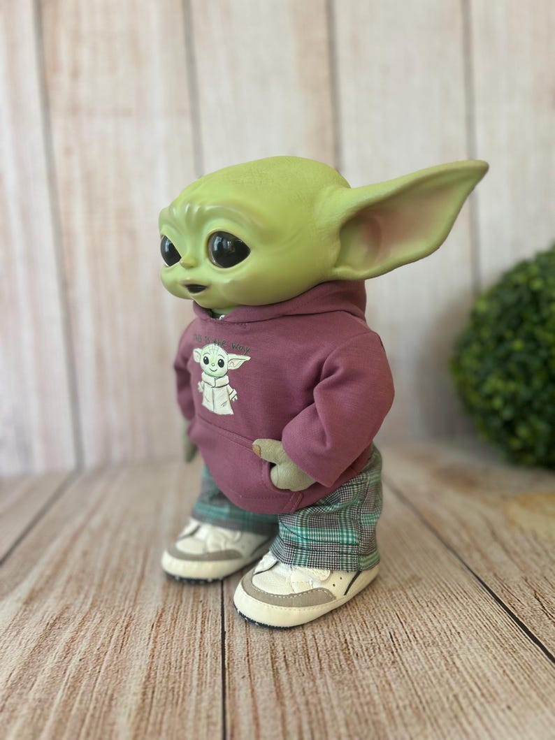 Op de afbeelding: Een groen wezenfiguurtje met grote donkere ogen en puntige oren, gekleed in een mauve hoodie met een kleine afbeelding van hetzelfde wezen op de voorkant, gecombineerd met een groen-zwarte geruite broek en wit-beige sneakers.
