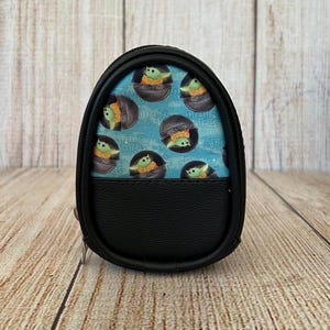 May include: A small, oval-shaped black zippered pouch featuring a light blue panel printed with a repeating pattern of Grogu from Star Wars inside floating pods. The lower half of the pouch is made of solid black textured material with a zipper closure.