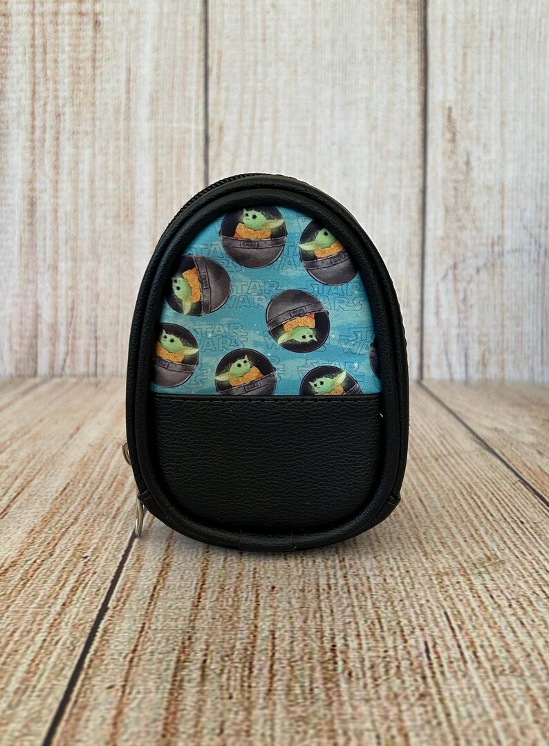 May include: A small, oval-shaped black zippered pouch featuring a light blue panel printed with a repeating pattern of Grogu from Star Wars inside floating pods. The lower half of the pouch is made of solid black textured material with a zipper closure.