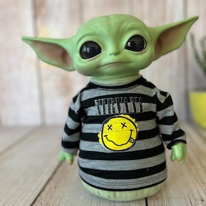 Peut inclure: Une poup&eacute;e verte avec de grands yeux noirs et de grandes oreilles. La poup&eacute;e porte une chemise &agrave; manches longues ray&eacute;e noire et grise avec le mot "NIRVANA" imprim&eacute; en haut et un smiley jaune avec un "X" dans chaque &oelig;il.