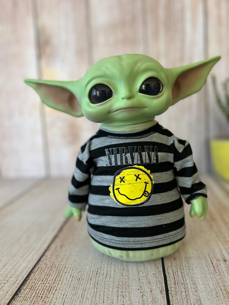 Peut inclure: Une poup&eacute;e verte avec de grands yeux noirs et de grandes oreilles. La poup&eacute;e porte une chemise &agrave; manches longues ray&eacute;e noire et grise avec le mot "NIRVANA" imprim&eacute; en haut et un smiley jaune avec un "X" dans chaque &oelig;il.