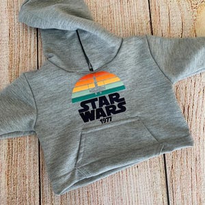 May include: A gray hooded sweatshirt with a front pocket. The sweatshirt features a colorful graphic of a sunset with a spaceship and the words "STAR WARS 1977" in black.