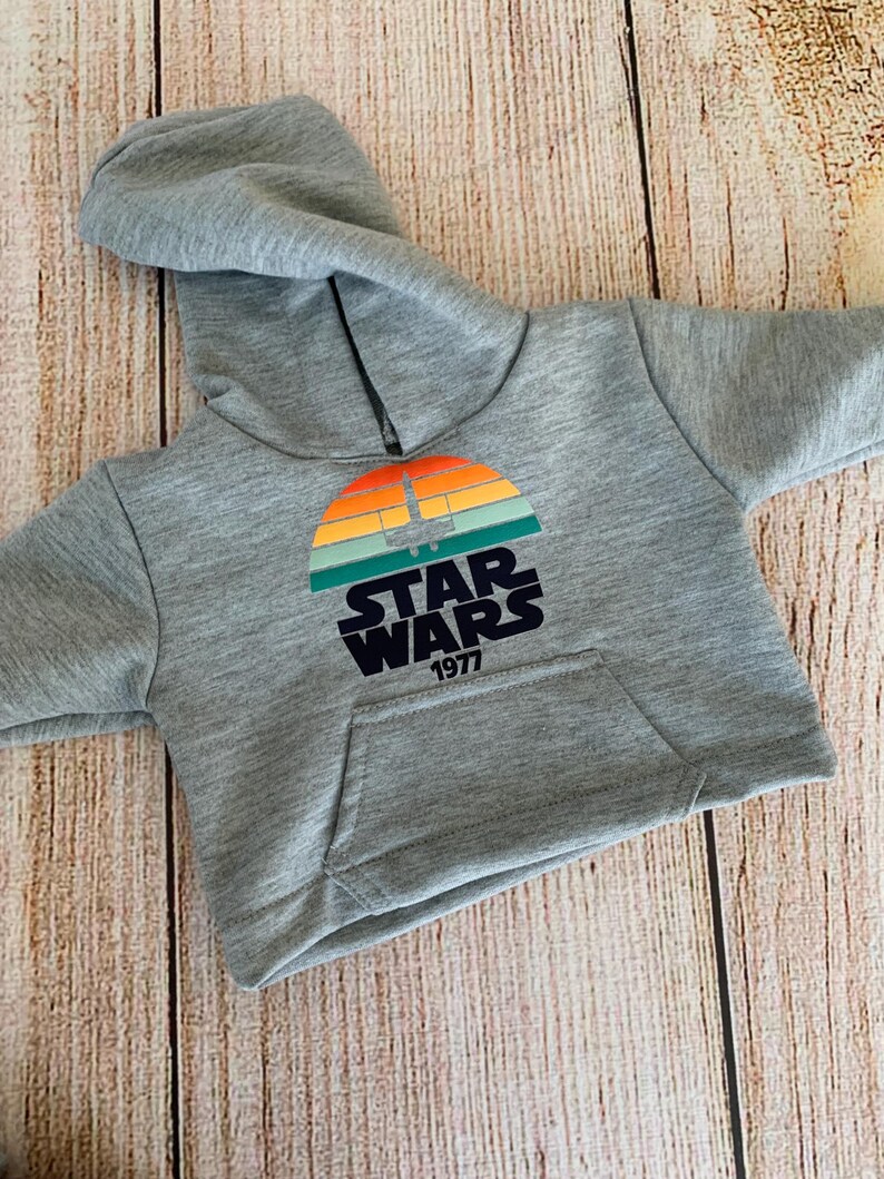May include: A gray hooded sweatshirt with a front pocket. The sweatshirt features a colorful graphic of a sunset with a spaceship and the words "STAR WARS 1977" in black.