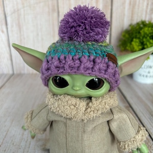 Op de afbeelding: Een groene pop met een paarse, turquoise en groene gehaakte muts met een paarse pompon. De pop draagt ook een beige jas met een donzige kraag en manchetten. De pop heeft grote zwarte ogen en puntige oren.