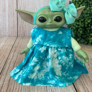 Puede incluir: Una muñeca de criatura verde con grandes ojos oscuros que lleva un vestido sin mangas con un patrón de teñido anudado en color verde azulado y blanco. La muñeca tiene una diadema a juego con un lazo y grandes orejas puntiagudas.