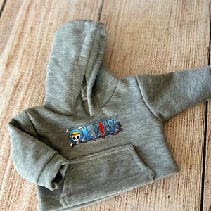 Op de afbeelding: Een grijs gem&ecirc;leerde hoodie met lange mouwen, voorzien van een kangoeroezak aan de voorzijde en een grafische print van het One Piece-logo, inclusief een schedel met strooien hoed en een rood silhouet op de tekst.