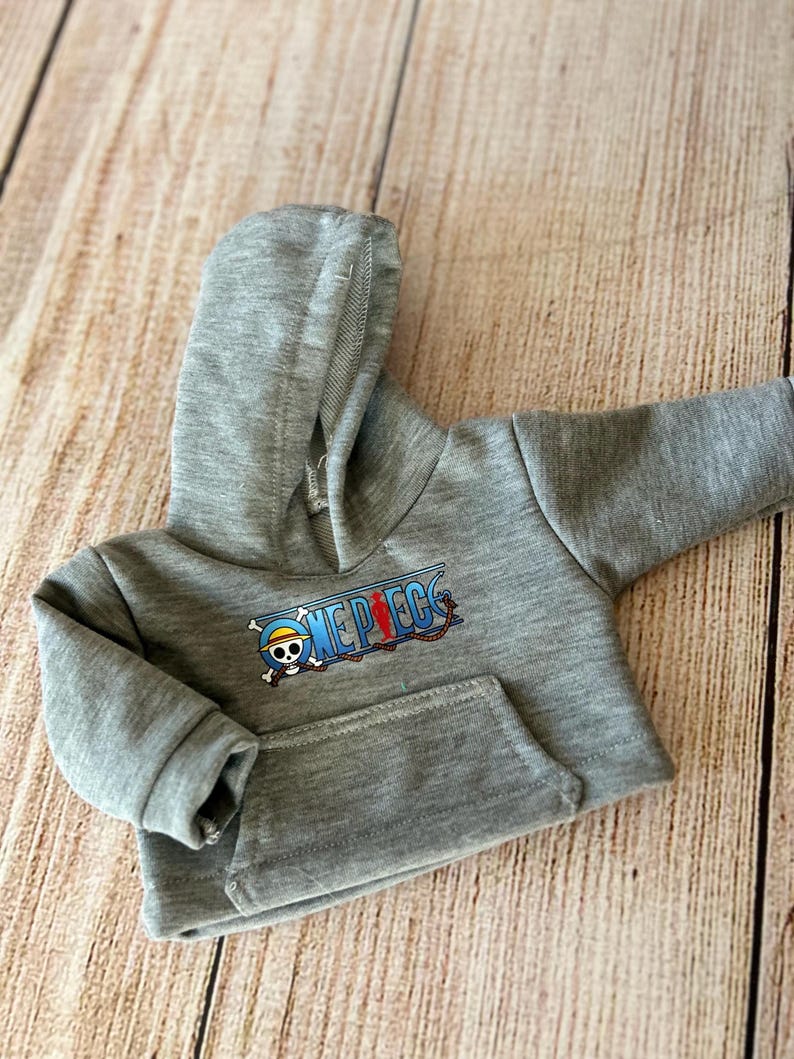 Op de afbeelding: Een grijs gem&ecirc;leerde hoodie met lange mouwen, voorzien van een kangoeroezak aan de voorzijde en een grafische print van het One Piece-logo, inclusief een schedel met strooien hoed en een rood silhouet op de tekst.