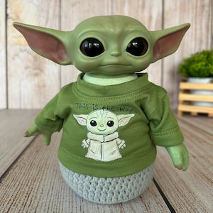 Puede incluir: Una figura verde de un personaje con orejas y ojos grandes, que lleva una camisa verde con el texto "This is the way" y un gráfico del personaje. La base es un diseño de ganchillo gris. La figura está sobre una superficie de madera.