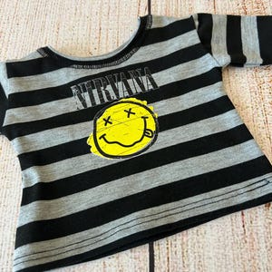 Peut inclure: Un t-shirt &agrave; rayures grises et noires avec le mot "NIRVANA" au-dessus d'un smiley jaune avec des yeux barr&eacute;s et une langue tir&eacute;e. Le t-shirt a un col et des manches noirs.