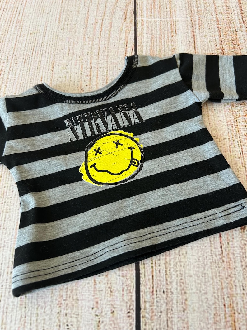 Peut inclure: Un t-shirt &agrave; rayures grises et noires avec le mot "NIRVANA" au-dessus d'un smiley jaune avec des yeux barr&eacute;s et une langue tir&eacute;e. Le t-shirt a un col et des manches noirs.
