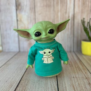 Peut inclure: Poup&eacute;e verte repr&eacute;sentant un personnage avec de grands yeux noirs et des oreilles pointues, portant un t-shirt turquoise &agrave; manches longues. Le t-shirt pr&eacute;sente une illustration blanche du personnage et le texte "This is the way."