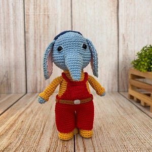 Puede incluir: Muñeco de elefante tejido a mano con cabeza azul, orejas rosas y ojos negros. Viste una camisa amarilla y un mono rojo con cinturón marrón. El muñeco mide aproximadamente 20 cm de altura.