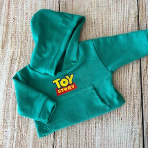 Op de afbeelding: Een felgroene hoodie met lange mouwen, voorzien van een kangoeroezak aan de voorzijde en het Toy Story-logo in geel en rood op de borst. Het kledingstuk heeft geribbelde boorden aan de mouwen en een capuchon met een zichtbare voering.