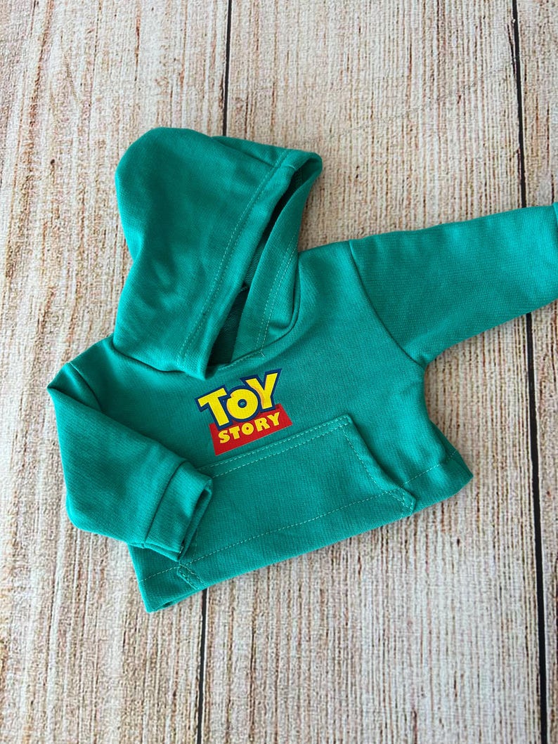 Op de afbeelding: Een felgroene hoodie met lange mouwen, voorzien van een kangoeroezak aan de voorzijde en het Toy Story-logo in geel en rood op de borst. Het kledingstuk heeft geribbelde boorden aan de mouwen en een capuchon met een zichtbare voering.