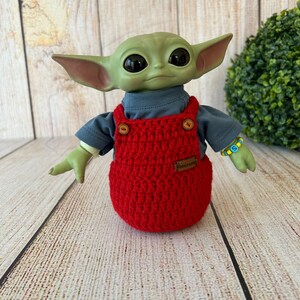 Op de afbeelding: Een groenhuidige pop met grote zwarte ogen, gekleed in een grijs shirt en een rode gehaakte overall. De pop heeft een kleine bruine patch op de overall en een kralenarmband. De pop staat op een houten oppervlak.