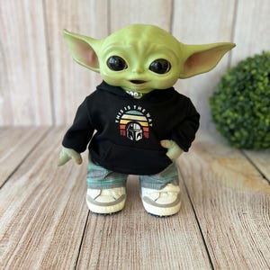 Op de afbeelding: Een groen wezenfiguurtje met grote zwarte ogen en puntige oren, gekleed in een zwarte hoodie met een grafische print van een helm en de tekst THIS IS THE WAY, gecombineerd met een groen-grijze geruite broek en witte sneakers.