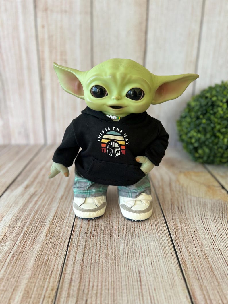 Op de afbeelding: Een groen wezenfiguurtje met grote zwarte ogen en puntige oren, gekleed in een zwarte hoodie met een grafische print van een helm en de tekst THIS IS THE WAY, gecombineerd met een groen-grijze geruite broek en witte sneakers.