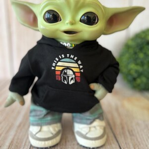 Op de afbeelding: Een groen wezenfiguur met grote zwarte ogen draagt een zwarte hoodie met een afbeelding van een helm en de tekst This Is The Way, gecombineerd met een grijze geruite broek en witte sneakers met grijze accenten op de zolen en neuzen.