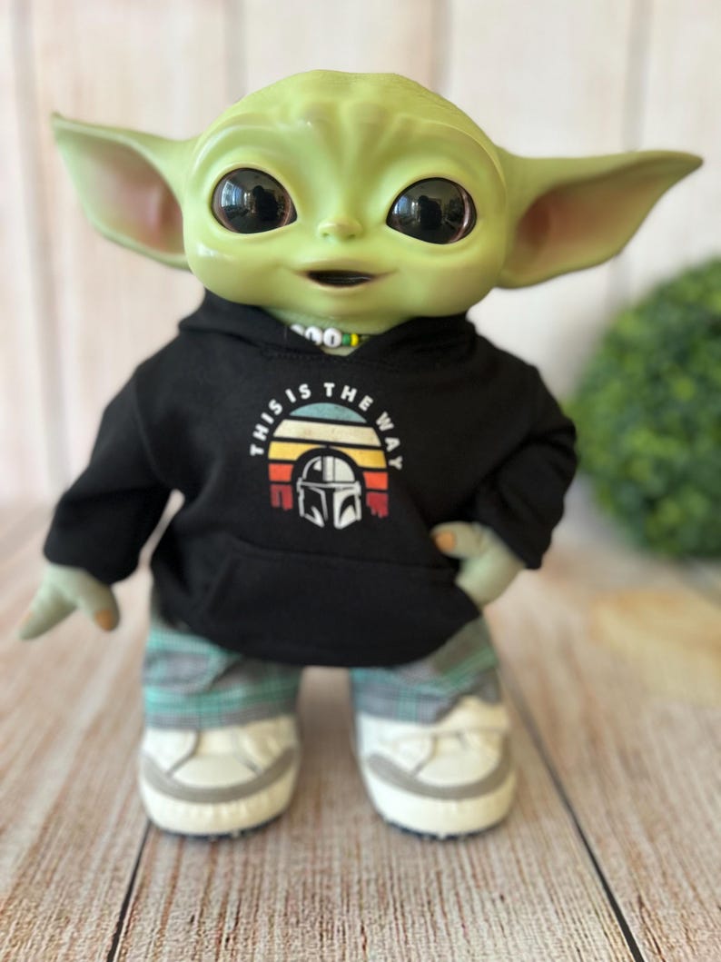 Op de afbeelding: Een groen wezenfiguur met grote zwarte ogen draagt een zwarte hoodie met een afbeelding van een helm en de tekst This Is The Way, gecombineerd met een grijze geruite broek en witte sneakers met grijze accenten op de zolen en neuzen.