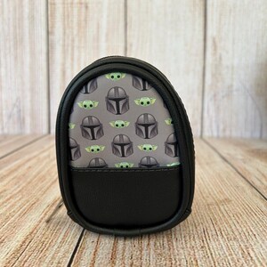 May include: A small, oval-shaped black zippered pouch featuring a repeating pattern of grey Mandalorian helmets and green baby alien characters on a grey background. The lower portion of the pouch is made of solid black material with a textured finish.