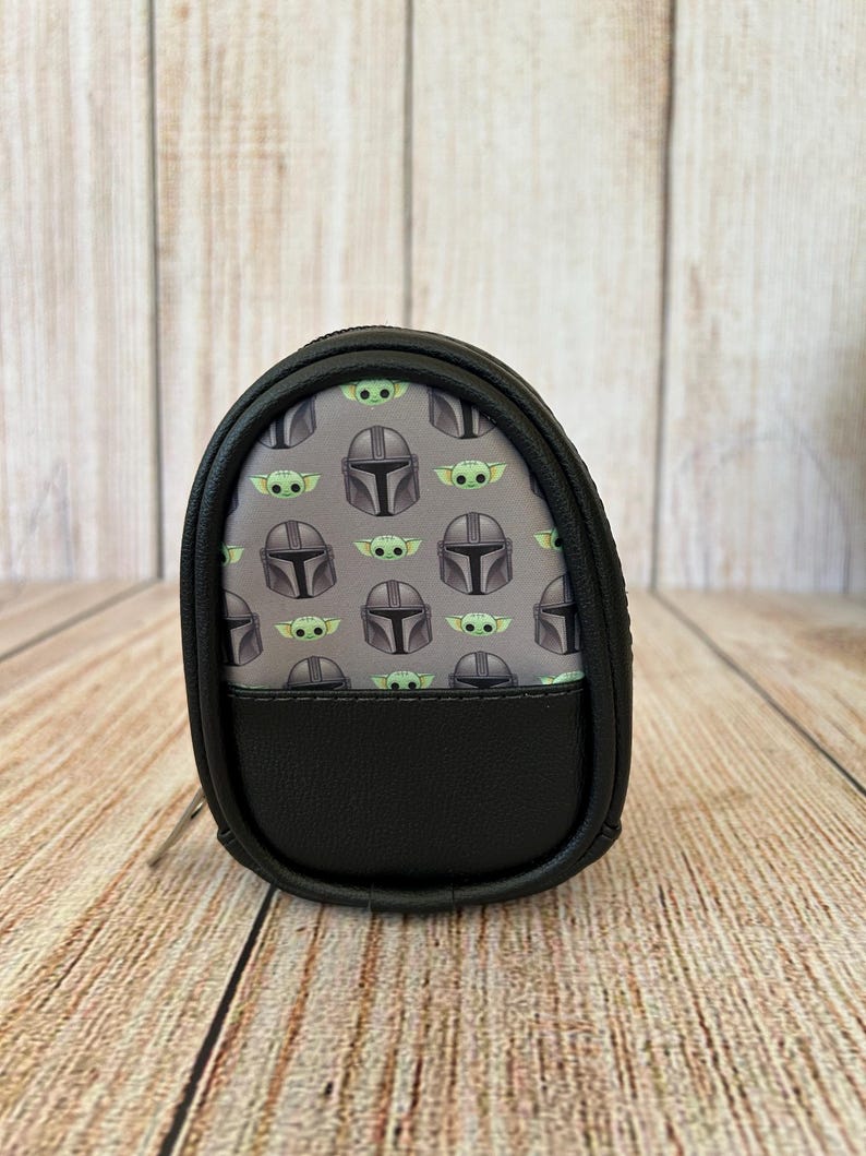 May include: A small, oval-shaped black zippered pouch featuring a repeating pattern of grey Mandalorian helmets and green baby alien characters on a grey background. The lower portion of the pouch is made of solid black material with a textured finish.