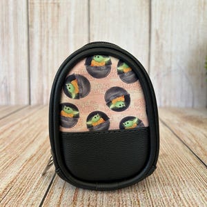 May include: A small, oval-shaped black zippered pouch featuring a patterned fabric panel with a repeating print of the character Grogu from Star Wars inside a floating pod. The pouch has a textured black faux leather lower section and black trim.