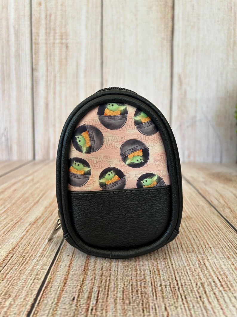 May include: A small, oval-shaped black zippered pouch featuring a patterned fabric panel with a repeating print of the character Grogu from Star Wars inside a floating pod. The pouch has a textured black faux leather lower section and black trim.