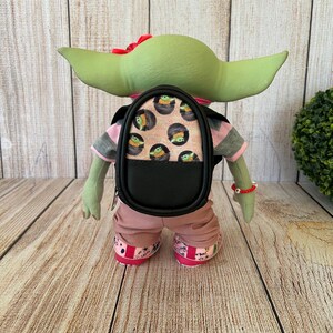 May include: A green creature figurine wearing a pink and gray striped shirt, pink pants, and pink patterned shoes. The figurine carries a small black backpack featuring a repeating print of a green character in a pod and a red bow on its head.