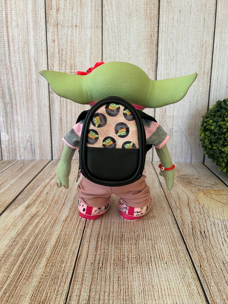 May include: A green creature figurine wearing a pink and gray striped shirt, pink pants, and pink patterned shoes. The figurine carries a small black backpack featuring a repeating print of a green character in a pod and a red bow on its head.