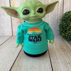 Peut inclure: Poup&eacute;e verte ressemblant &agrave; un personnage de Star Wars, portant un t-shirt &agrave; manches longues turquoise. Le t-shirt pr&eacute;sente un motif de coucher de soleil r&eacute;tro avec les mots "STAR WARS 1977". La poup&eacute;e a de grands yeux noirs et de grandes oreilles.