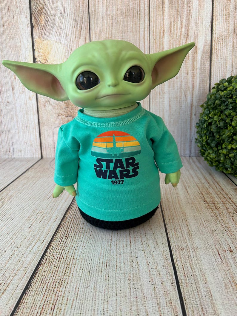 Peut inclure: Poup&eacute;e verte ressemblant &agrave; un personnage de Star Wars, portant un t-shirt &agrave; manches longues turquoise. Le t-shirt pr&eacute;sente un motif de coucher de soleil r&eacute;tro avec les mots "STAR WARS 1977". La poup&eacute;e a de grands yeux noirs et de grandes oreilles.