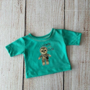 Peut inclure: Un t-shirt turquoise &agrave; manches courtes avec un motif brod&eacute; d'un personnage poilu avec une bandouli&egrave;re. Le texte "Chewie and Chill" est au-dessus du personnage. Le t-shirt est pr&eacute;sent&eacute; sur une surface en bois clair.