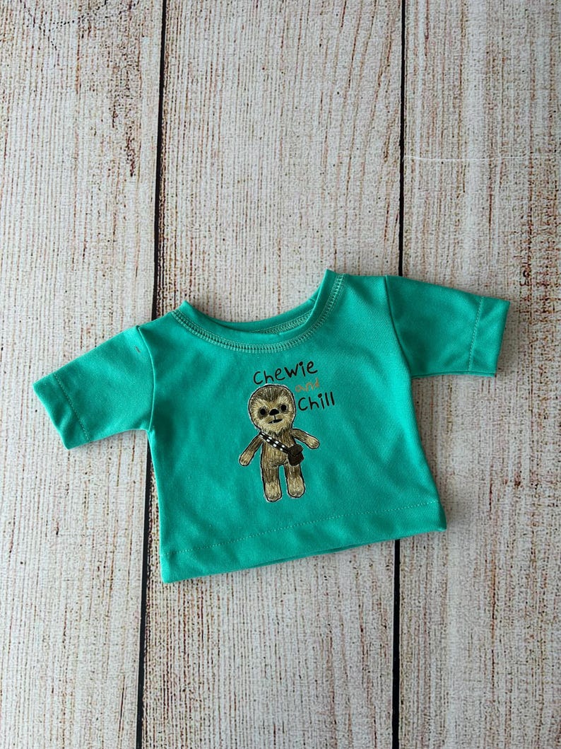 Peut inclure: Un t-shirt turquoise &agrave; manches courtes avec un motif brod&eacute; d'un personnage poilu avec une bandouli&egrave;re. Le texte "Chewie and Chill" est au-dessus du personnage. Le t-shirt est pr&eacute;sent&eacute; sur une surface en bois clair.