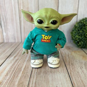 Op de afbeelding: Een groen wezenfiguurtje met grote zwarte ogen en puntige oren, gekleed in een blauwgroene hoodie met de tekst TOY STORY in geel en rood, een grijze broek met ruitjespatroon en witte sneakers met klittenbandsluiting aan de voeten.