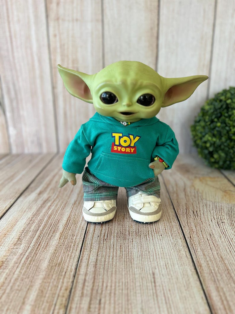 Op de afbeelding: Een groen wezenfiguurtje met grote zwarte ogen en puntige oren, gekleed in een blauwgroene hoodie met de tekst TOY STORY in geel en rood, een grijze broek met ruitjespatroon en witte sneakers met klittenbandsluiting aan de voeten.