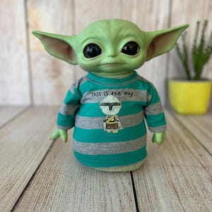 Peut inclure: Poup&eacute;e verte ressemblant &agrave; un personnage d'une s&eacute;rie de science-fiction populaire. La poup&eacute;e porte un t-shirt ray&eacute; turquoise et gris avec le texte "THIS IS THE WAY" et un graphique. La poup&eacute;e a de grands yeux noirs et des oreilles pointues.