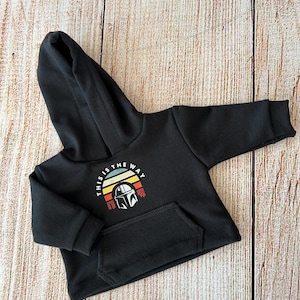 Op de afbeelding: Een zwarte hoodie met lange mouwen en een kangoeroezak aan de voorzijde. Op de borst staat een afbeelding van een helm in een halfrond zonsondergangontwerp met de tekst "THIS IS THE WAY" in witte hoofdletters erboven.