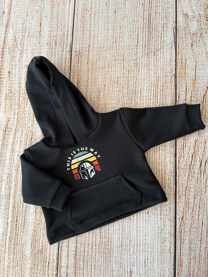 Op de afbeelding: Een zwarte hoodie met lange mouwen en een kangoeroezak aan de voorzijde. Op de borst staat een afbeelding van een helm in een halfrond zonsondergangontwerp met de tekst "THIS IS THE WAY" in witte hoofdletters erboven.