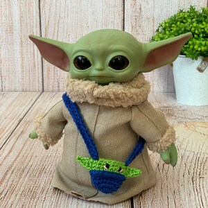 K&ouml;nnte beinhalten: Ein gr&uuml;nes Pl&uuml;schtier mit gro&szlig;en Augen und einem braunen Fellkragen, das eine beige Jacke mit einer blauen und gr&uuml;nen geh&auml;kelten Tasche tr&auml;gt.
