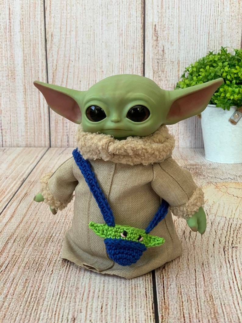 K&ouml;nnte beinhalten: Ein gr&uuml;nes Pl&uuml;schtier mit gro&szlig;en Augen und einem braunen Fellkragen, das eine beige Jacke mit einer blauen und gr&uuml;nen geh&auml;kelten Tasche tr&auml;gt.