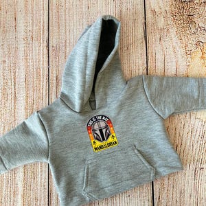 May include: A gray hooded sweatshirt with a kangaroo pocket. The front features a colorful patch with the text "THIS IS THE WAY" and a Mandalorian helmet design. The sweatshirt is displayed on a light brown wooden surface.