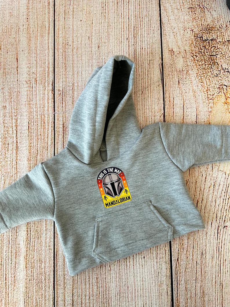 May include: A gray hooded sweatshirt with a kangaroo pocket. The front features a colorful patch with the text "THIS IS THE WAY" and a Mandalorian helmet design. The sweatshirt is displayed on a light brown wooden surface.