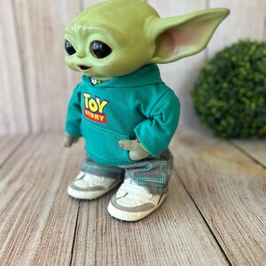 Op de afbeelding: Een groen wezenfiguurtje met grote zwarte ogen en puntige oren, gekleed in een blauwgroene hoodie met de tekst Toy Story op de voorkant, een grijze broek met ruitjespatroon en witte sneakers, weergegeven in een zijaanzicht.