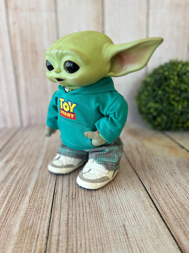 Op de afbeelding: Een groen wezenfiguurtje met grote zwarte ogen en puntige oren, gekleed in een blauwgroene hoodie met de tekst Toy Story op de voorkant, een grijze broek met ruitjespatroon en witte sneakers, weergegeven in een zijaanzicht.