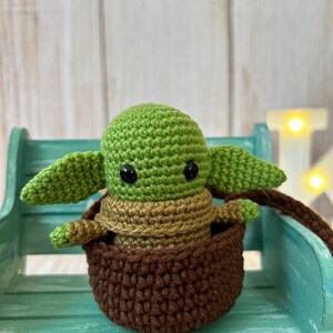 May include: A green crocheted baby Yoda doll sitting in a brown crocheted basket. The doll has large black eyes and is sitting on a teal bench.