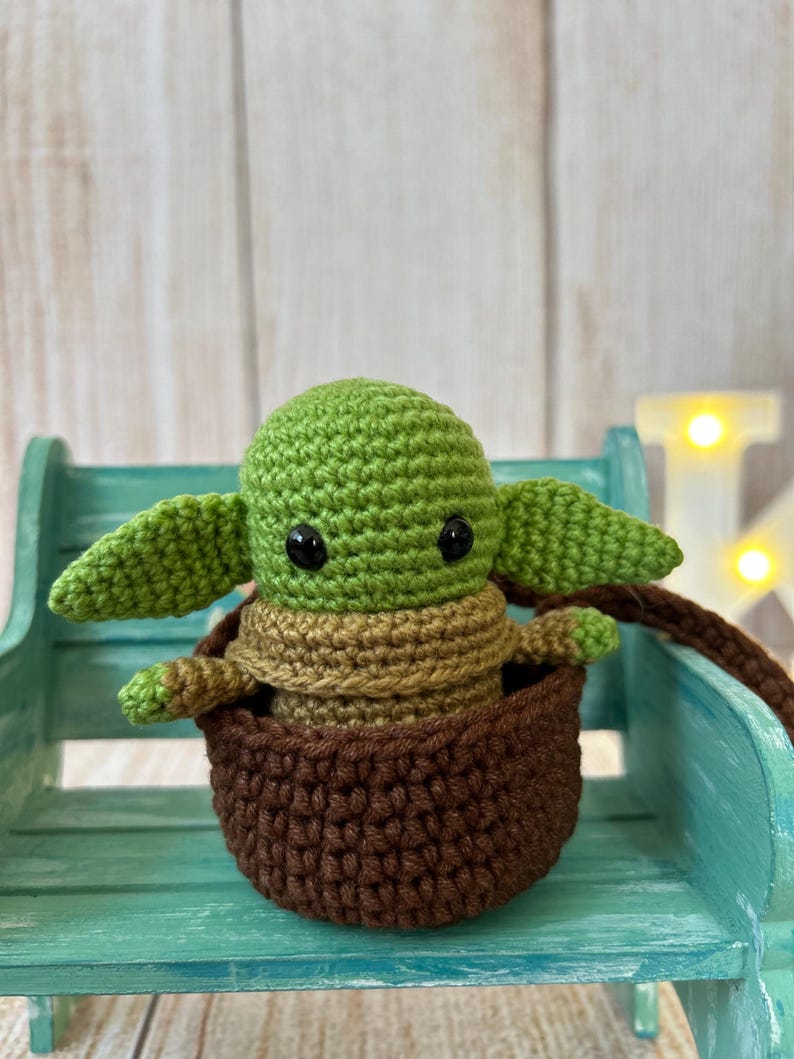 May include: A green crocheted baby Yoda doll sitting in a brown crocheted basket. The doll has large black eyes and is sitting on a teal bench.