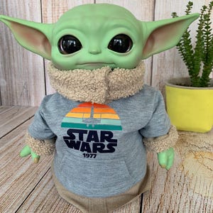 May include: A green-skinned doll with large black eyes, wearing a gray Star Wars hoodie with a retro sunset design and the text "STAR WARS 1977". The doll has large ears and a tan fuzzy collar and cuffs.