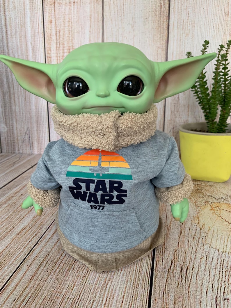 May include: A green-skinned doll with large black eyes, wearing a gray Star Wars hoodie with a retro sunset design and the text "STAR WARS 1977". The doll has large ears and a tan fuzzy collar and cuffs.